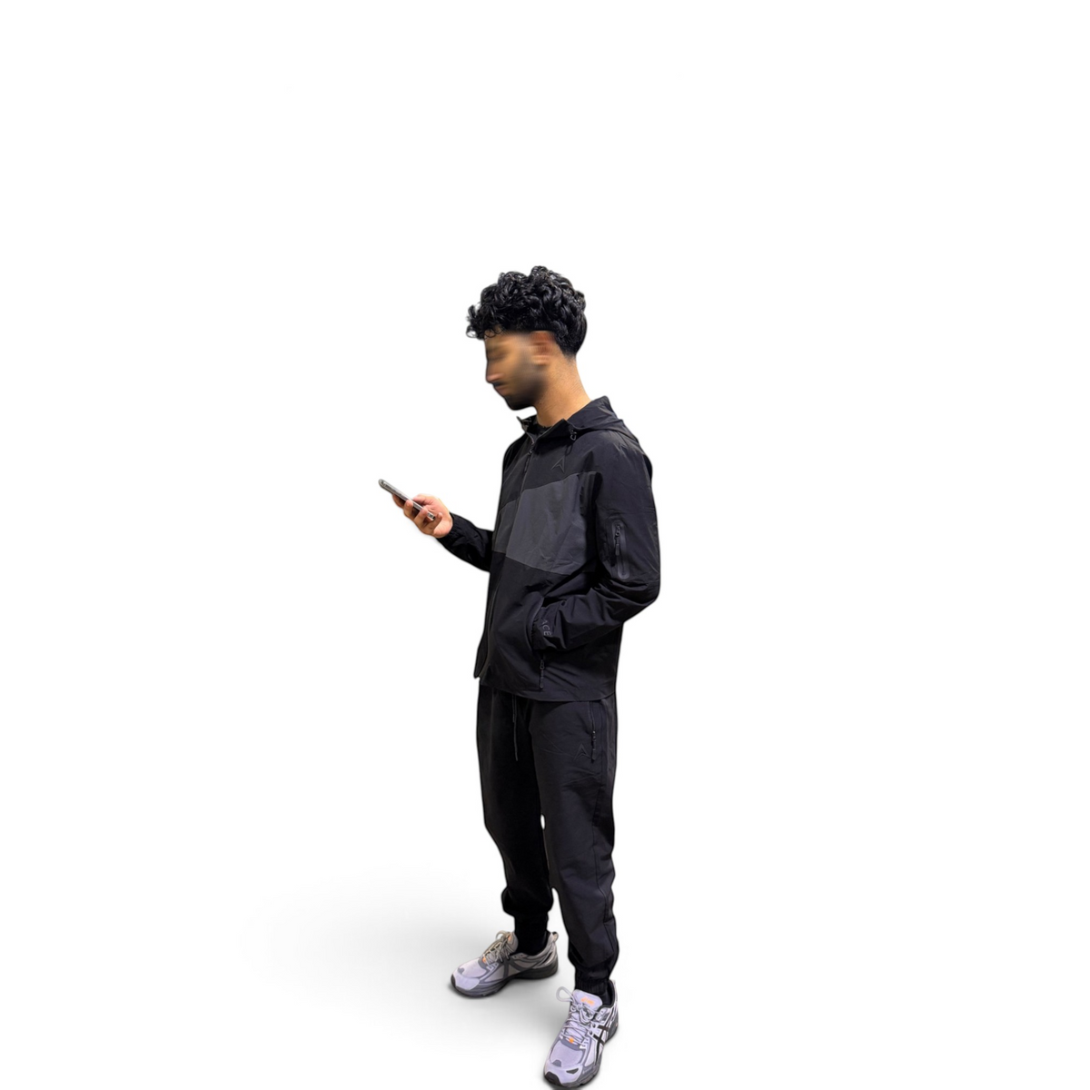 A man wearing a black jacket, black pants, and gray sneakers stands in profile against a solid white background, holding a smartphone.
