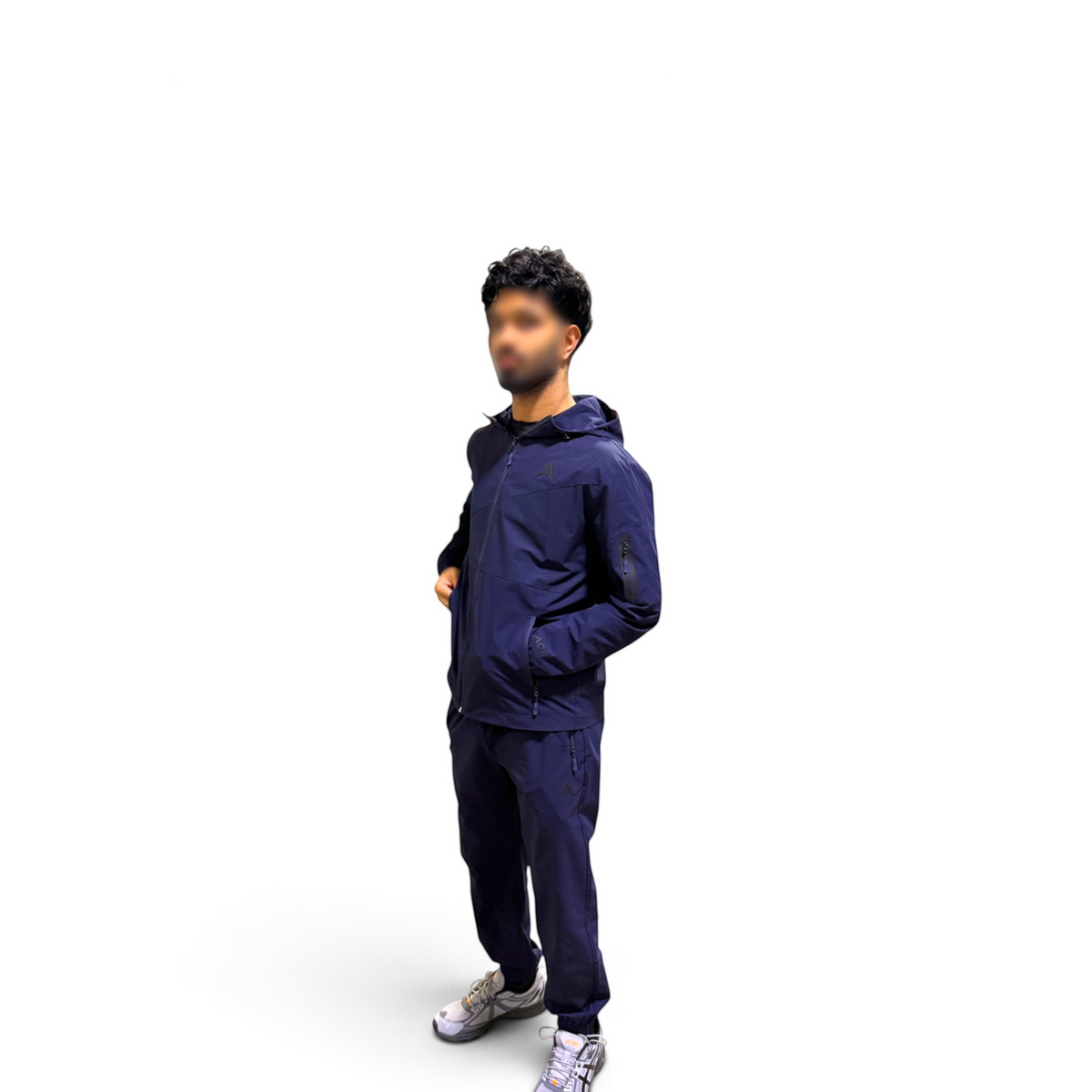 A man wearing a navy blue hooded jacket and matching navy pants, standing against a plain white background with his face blurred.