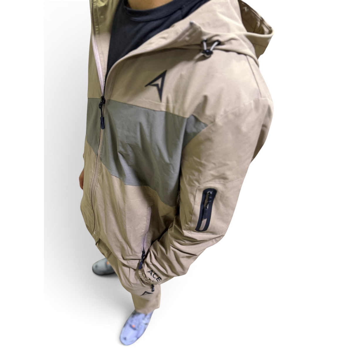 A person wearing a beige and gray color-blocked hooded jacket with black zipper details and a black logo on the chest, paired with a dark shirt and light blue patterned socks, against a plain white background.