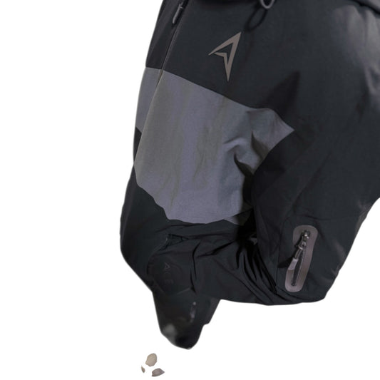Close-up of a black and gray outdoor jacket sleeve with a gray reflective-looking triangular logo on the upper chest and a zippered pocket on the lower arm, isolated on a white background.